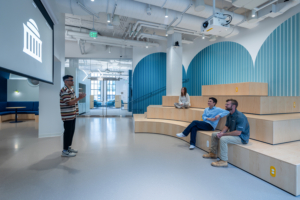 Photo of a four-tiered platform, where two people sit. Another person speaks to them in front of a screen.