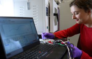 An MIT graduate student conducts an electrochemical experiment in her lab, monitoring results on a connected laptop.