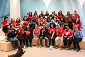 Photo of 29 people, seated in three rows, mostly wearing red shirts and holding signs of different MIT classes or with phrases like "happy first day of class." A dog is in the foreground.