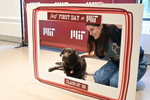 Winni the dog poses with a student inside an oversized frame that says "Last first day at MIT, Class of 2026"