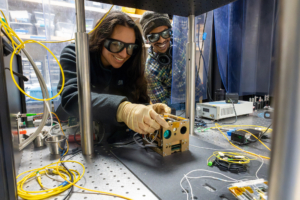 Two researchers wearing safety glasses work together in a laboratory setting. The person in front, wearing a black jacket and light gloves, holds a tan-colored electronic device while examining it closely. Behind them, a colleague in a plaid shirt and beanie smiles at the camera. The lab is equipped with yellow networking cables, testing equipment, and various electronic instruments, with city buildings visible through the windows in the background.