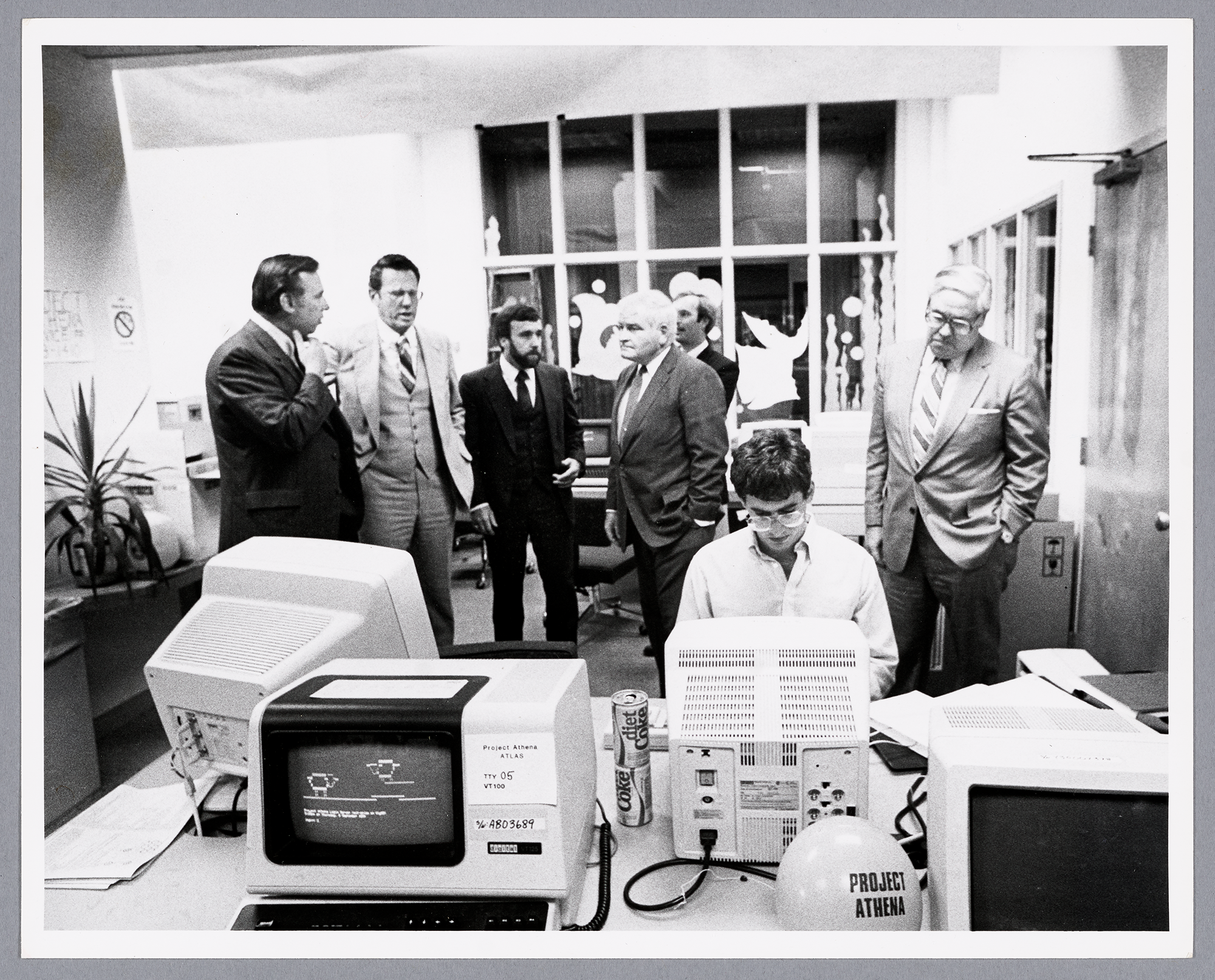 black and white photo of a student working at a computer while an older man in a suit looks on. There are multiple computer monitors on the desk, a balloon that says 