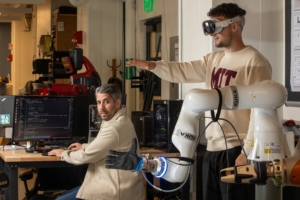 Two researchers, one seated at a keyboard, one standing and wearing VR goggles to interact with a robotic arm